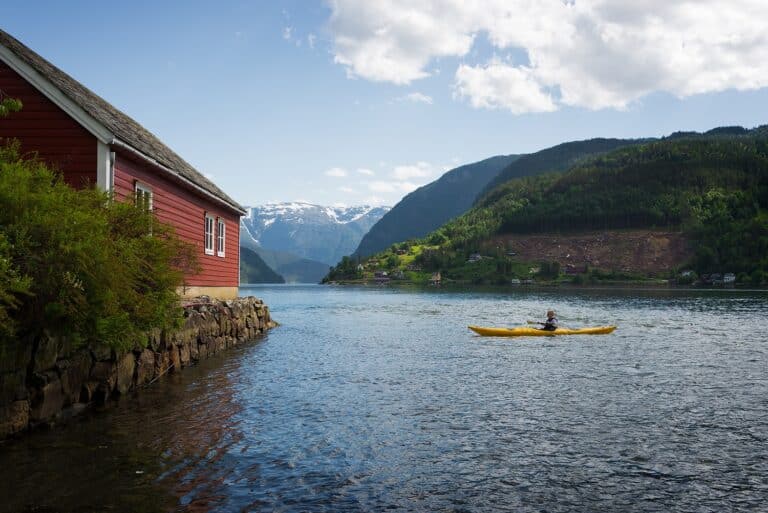 Iemand in een gele kajak op het Hardangerfjord, naast een rood houten huisje
