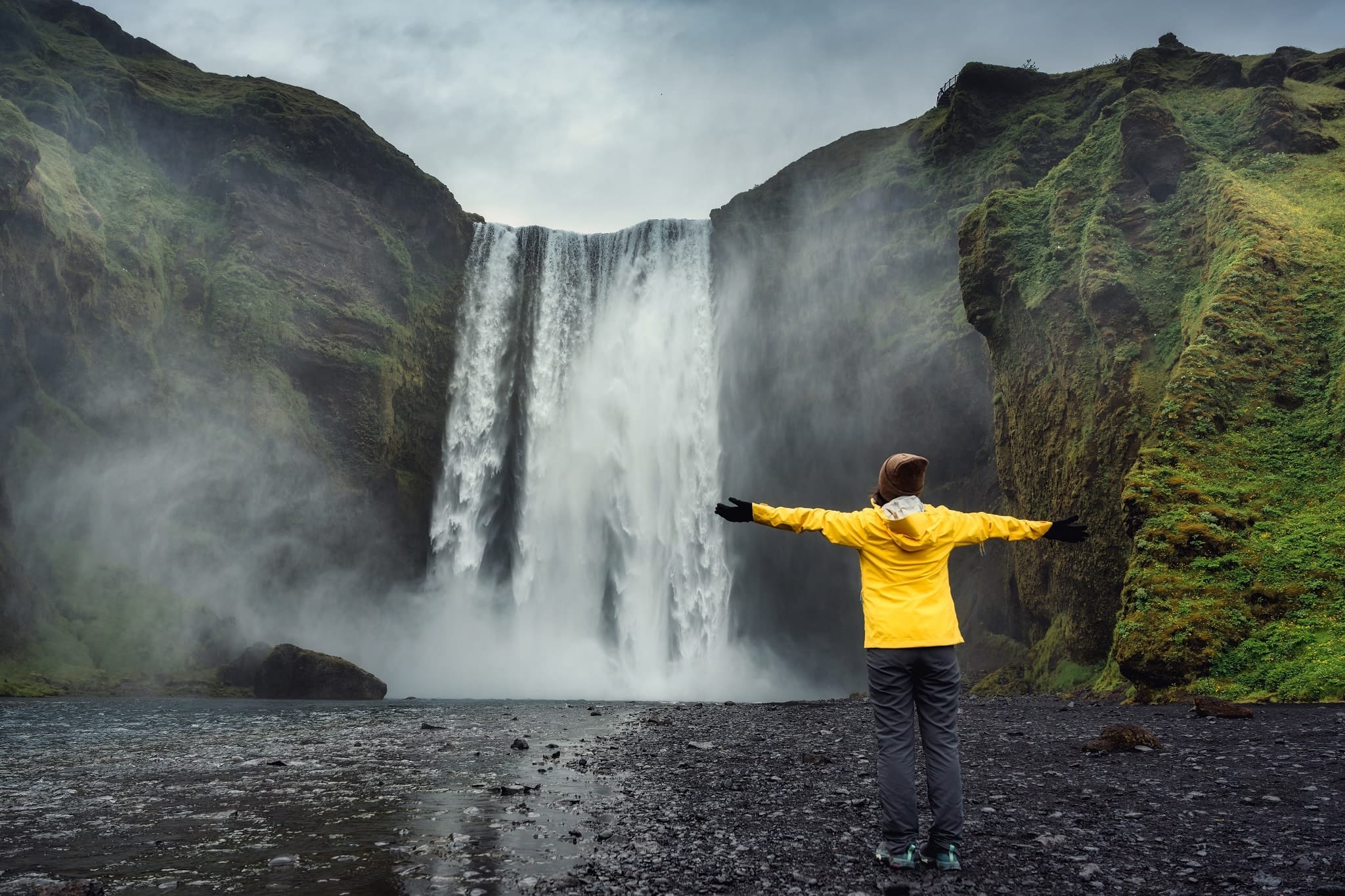 Vrouw in gele jas staat met armen wijd voor Skogafoss waterval op IJsland