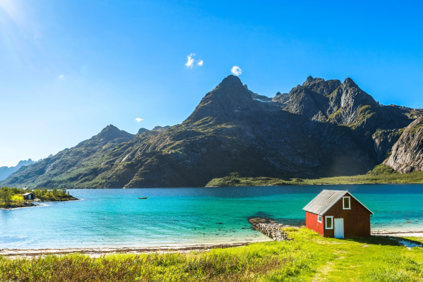 Rood houten huisje op het strand, aan helder blauw water. Op de achtergrond bergen.